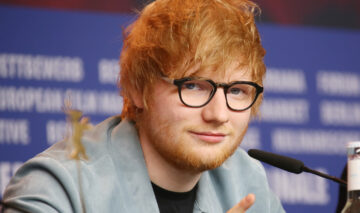 Ed Sheeran la o conferință de presă purtând ochelari rotuni negri, un tricou negru și un sacou gri.