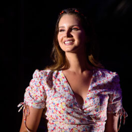 Lana del Rey zâmbește pe o scenă. Este îmbrăcată cu o rochie înflorată cu mânecă scurtă și are părul lăsat pe spate. Are unul dintre cele mai bune albume de vară din toate timpurile.