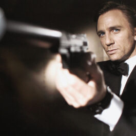 James Bond cu un pistol în mână. Este îmbrăcat în celebrul lui costum elegant cu papion și cu un ceas mare la mână. Ne putem imagina cum pe fundal cântă una dintre cele mai bune melodii din filmele cu James Bond
