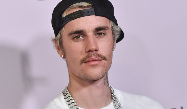 Justin Bieber pe covorul roșu purtând un tricou alb, cu un lanț argintiu la gât și cu o lapcă neagră pe cap întoarsă cu cozorocul la spate