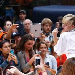 Miley Cyrus în mulțime cântând piese pentru karaoke. Este îmbrăcată cu un top alb și are pe buze ruj roșu foarte aprins.