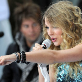 Taylor Swift în concert. Este îmbrăcată cu o rochie strălucitoare și ține în mână un microfon. Acum Taylor Swift își reface albumele.