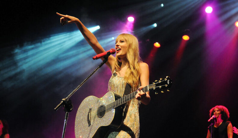 Taylor Swift este pe scenă, cu chitara în mână. Poartă o rochie aurie, mulată și lungă care îi scoate în evidență trupul. Părul este lăsat pe spate și puțin ondulat.