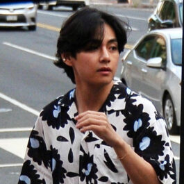 V de la BTS, fotografiat pe strada, intr-o camasa alba cu flori negre, cu parul aranjat cu carare pe mijloc