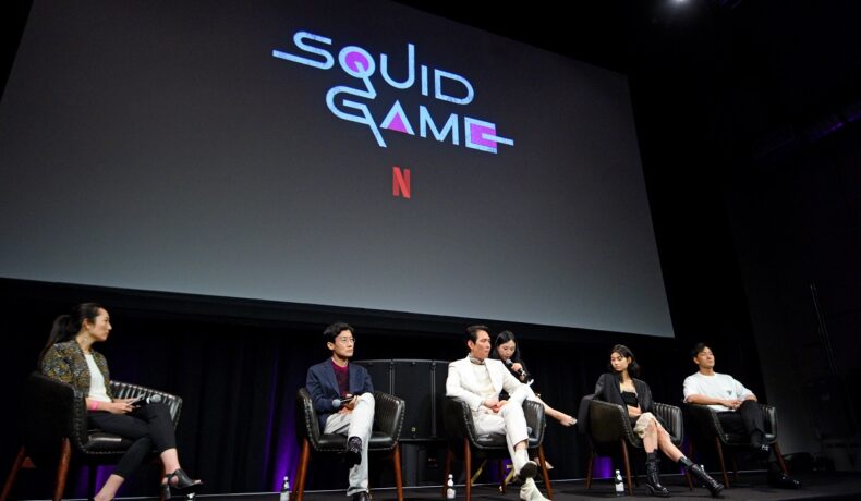 Actorii din primul sezon Squid Game cu numele emisiunii deasupra lor pe un ecran imens