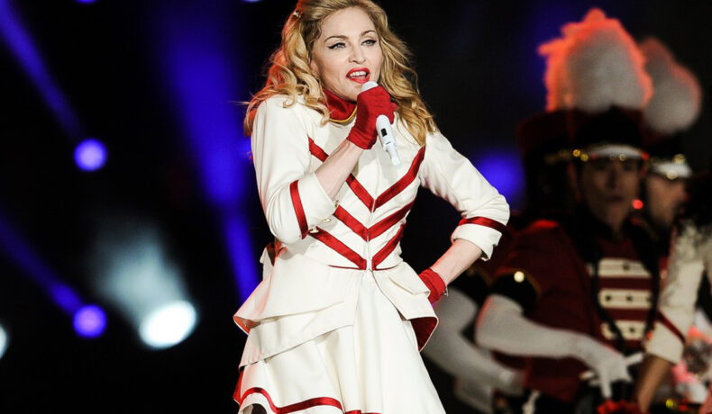 Madonna începe turneul pe care fanii îl așteaptă cu nerăbdare. Cântăreața este pe scenă cu o rochie albă cu bungi roșii și mănuși asortate