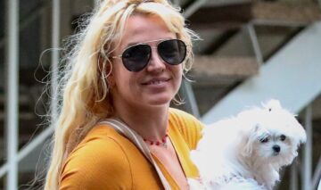 Britney Spears îmbrăcată într-o rochie portocalie purtând ochelari de soare și ținând un cățel alb în brațe
