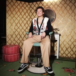 Harry Styles îmbrăcat casual în timp ce stă pe un scaun și zâmbește larg