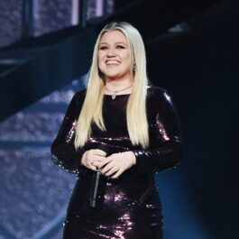 Kelly Clarkson îmbrăcată în negru în timp ce ține microfonul în mână și zâmbește
