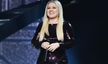 Kelly Clarkson îmbrăcată în negru în timp ce ține microfonul în mână și zâmbește