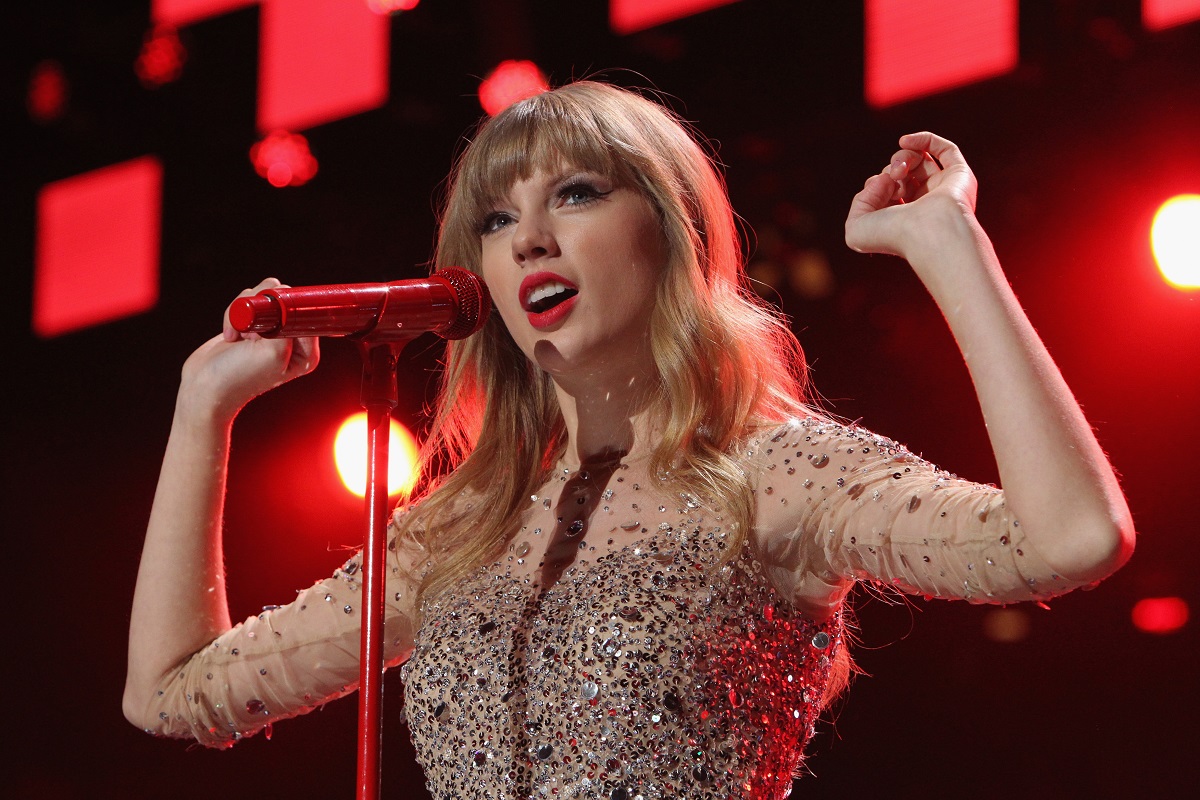 Taylor Swift îmbrăcată într-o rochie strălucitoare, cu un fundal roșu în spate, în timp ce cântă