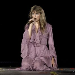Taylor Swift îmbrăcată într-o rochie mov în timp ce stă pe genunchi și cântă la microfon