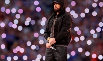 Eminem îmbrăcat cu un hanorac negru și o șapcă în timp ce ține microfonul în mână