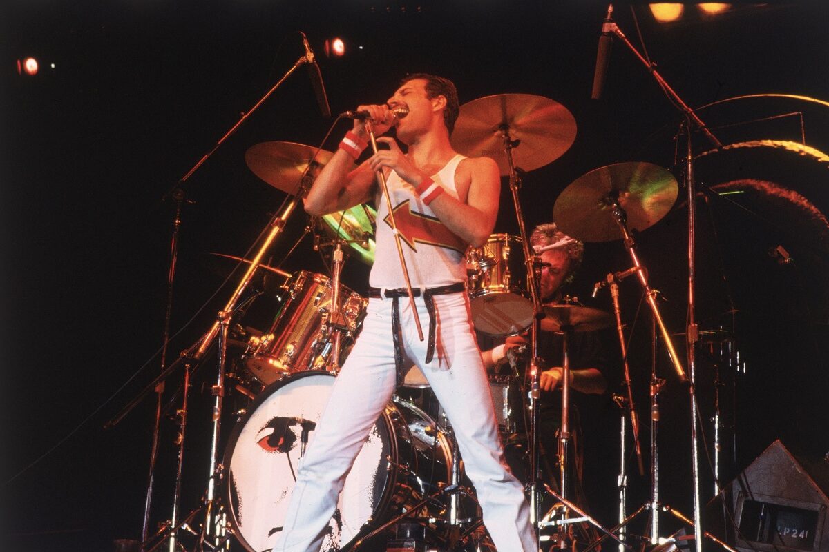 Freddie Mercury îmbrăcat cu o pereche de blugi deschiși la culoare și un maiou alb în timp ce stă în fața unui set de tobe și cântă la microfon