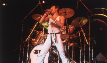Freddie Mercury îmbrăcat cu o pereche de blugi deschiși la culoare și un maiou alb în timp ce stă în fața unui set de tobe și cântă la microfon