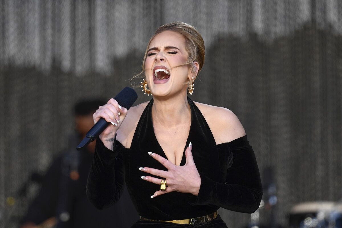 Adele îmbrăcată cu o rochie neagră decupată în zona umerilor în timp ce cântă la microfon