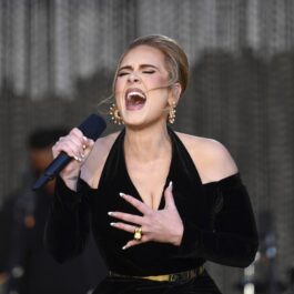 Adele îmbrăcată cu o rochie neagră decupată în zona umerilor în timp ce cântă la microfon