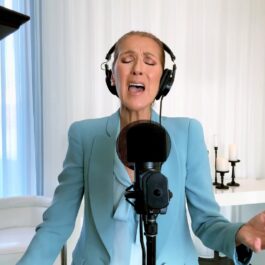 Celine Dion îmbrăcată într-un costum albastru deschis cu o pereche de căști pe cap în timp ce cântă la un microfon fix