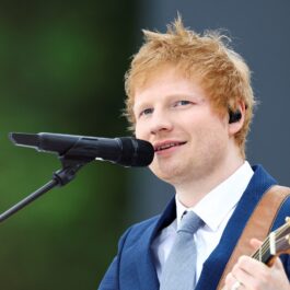 Ed Sheeran îmbrăcat într-un costum albastru în timp ce cântă la chitară și cu vocea la un microfon