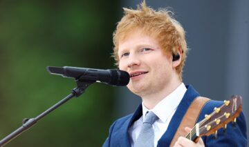 Ed Sheeran îmbrăcat într-un costum albastru în timp ce cântă la chitară și cu vocea la un microfon
