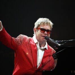 Elton John îmbrăcat într-un sacou roșu și ochelari de soare în timp ce cântă la microfon