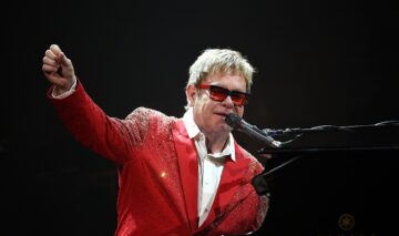 Elton John îmbrăcat într-un sacou roșu și ochelari de soare în timp ce cântă la microfon