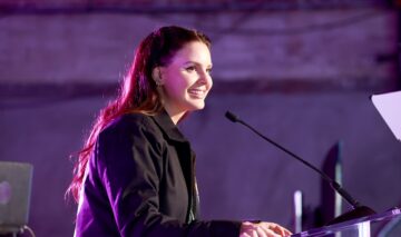 Lana Del Rey îmbrăcată într-o jachetă neagră în timp ce vorbește la microfon și zâmbește