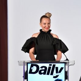 Sia îmbrăcată într-o rochie neagră cu umeri goi în timp ce ține un discurs