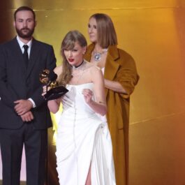 Taylor Swift îmbrăcată într-o rochie albă în timp ce își revendică premiul Grammy și Celine Dion îmbrăcată într-o ținută portocalie în spatele ei