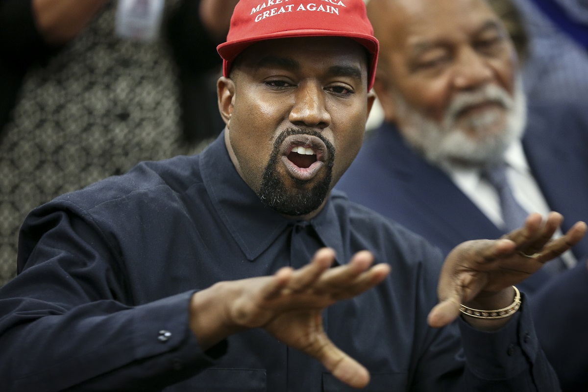 Kanye West îmbrăcat cu o cămașă neagră și o șapcă roșie vorbin și gesticulând