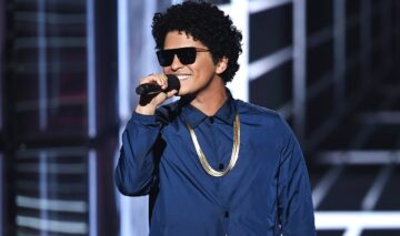 Bruno Mars îmbrăcat cu o geacă albastră, un lanț strălucitor și ochelari de soare în timp ce zâmbește și cântă la microfon