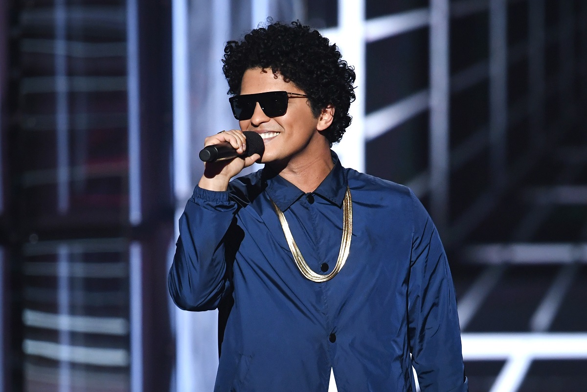 Bruno Mars îmbrăcat cu o geacă albastră, un lanț strălucitor și ochelari de soare în timp ce zâmbește și cântă la microfon