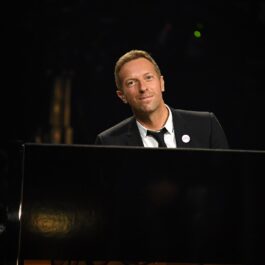 Chris Martin îmbrăcat elegant într-un costum negru cu cămașă albă în timp ce cântă la pian