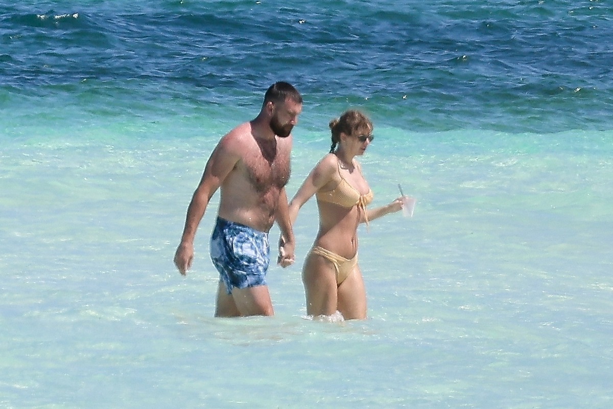 Taylor Swift și Travis Kelce îmbrăcați în costume de baie și ținându-se de mână în timp ce se bălăcesc în apă