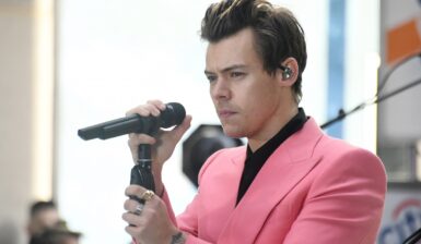 Harry Styles îmbrăcat cu o cămașă neagră pe sub un sacou roz în timp ce ține un microfon în mâna dreaptă