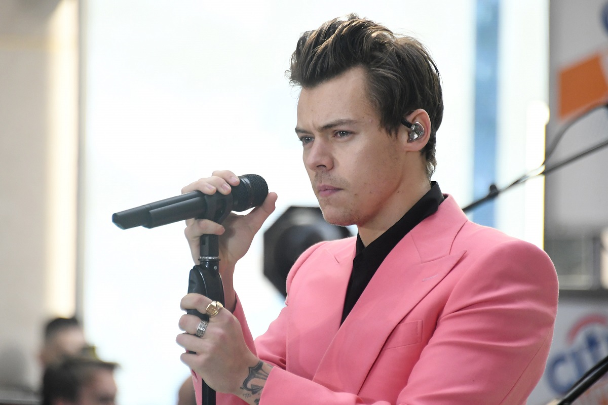 Harry Styles îmbrăcat cu o cămașă neagră pe sub un sacou roz în timp ce ține un microfon în mâna dreaptă