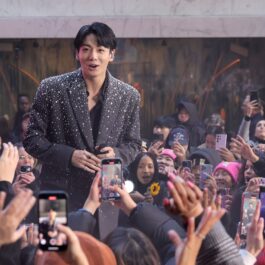 Jungkook îmbrăcat într-un sacou gri cu pietre strălucitoare în timp ce este înconjurat de o mulțime de fani care îl filmează