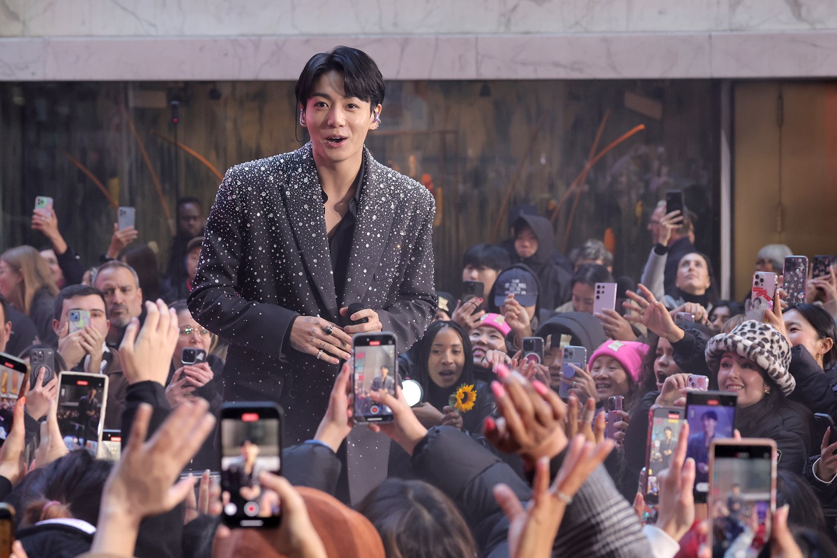 Jungkook îmbrăcat într-un sacou gri cu pietre strălucitoare în timp ce este înconjurat de o mulțime de fani care îl filmează