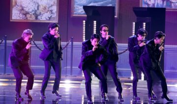 Membrii BTS îmbrăcați în costume negre în timp ce cântă și dansează pe scenă