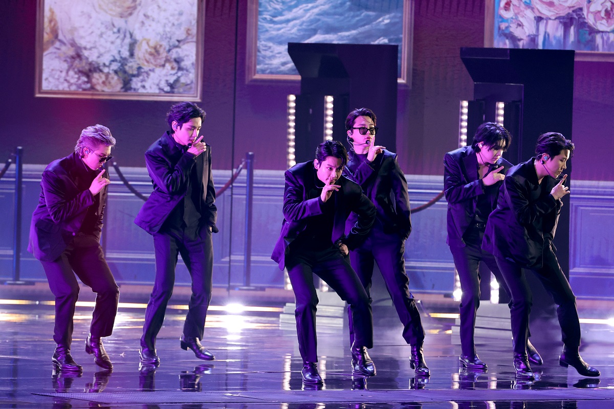 Membrii BTS îmbrăcați în costume negre în timp ce cântă și dansează pe scenă