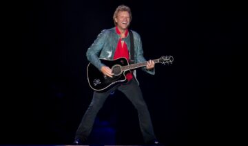 Jon Bon Jovi îmbrăcat într-o ținută casual în timp ce cântă la o chitară neagră și zâmbește larg