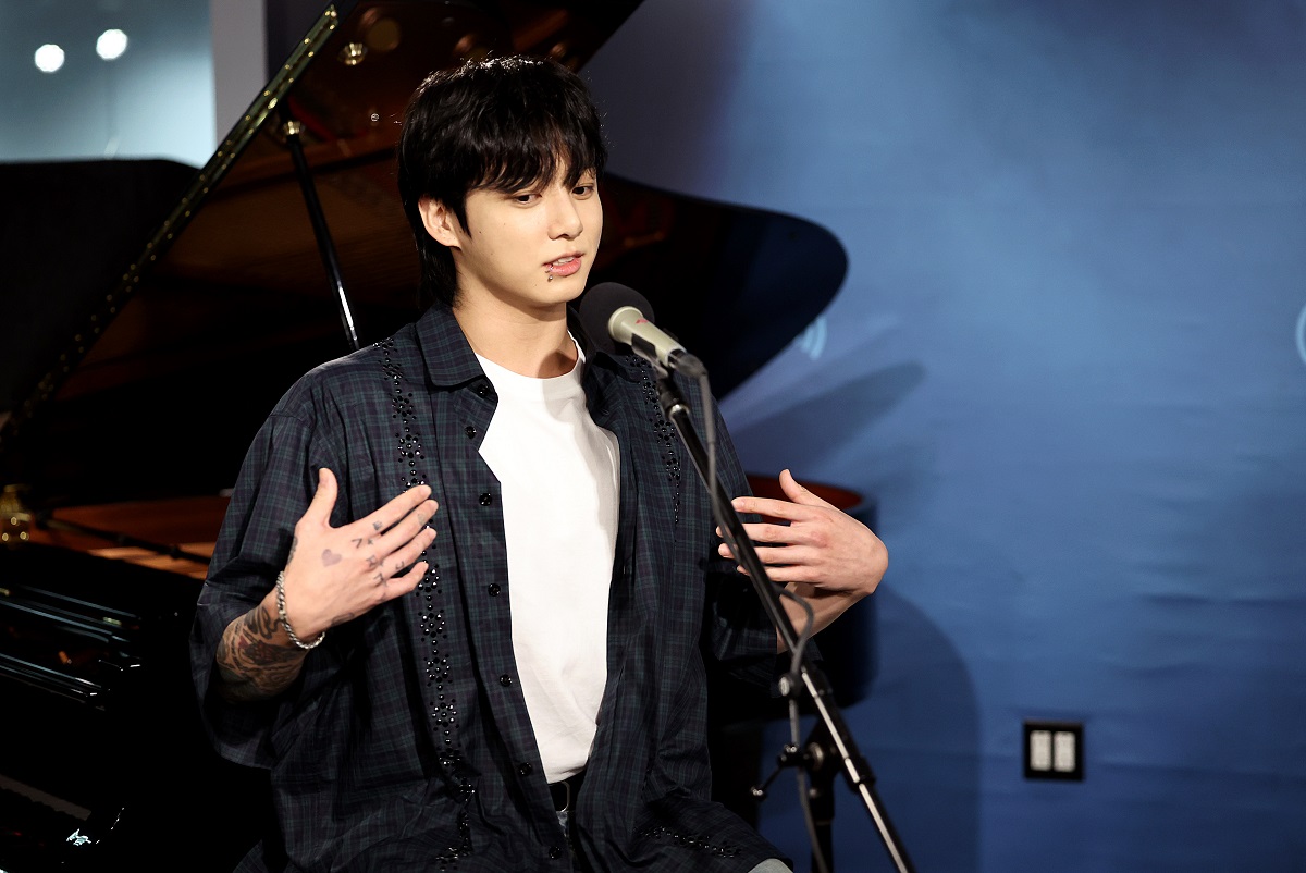 Jungkook îmbrăcat cu un tricou alb sub o cămașă neagră în carouri în timp ce vorbește la microfon