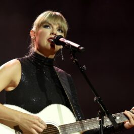Taylor Swift îmbrăcată într-o rochie neagră cu umerii decupați în timp ce cântă la microfon și la o chitarp albă