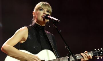 Taylor Swift îmbrăcată într-o rochie neagră cu umerii decupați în timp ce cântă la microfon și la o chitarp albă