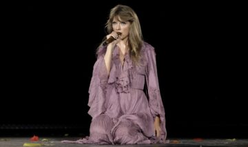 Taylor Swift îmbrăcată într-o rochie mov în timp ce stă în genunchi și cântă la microfon