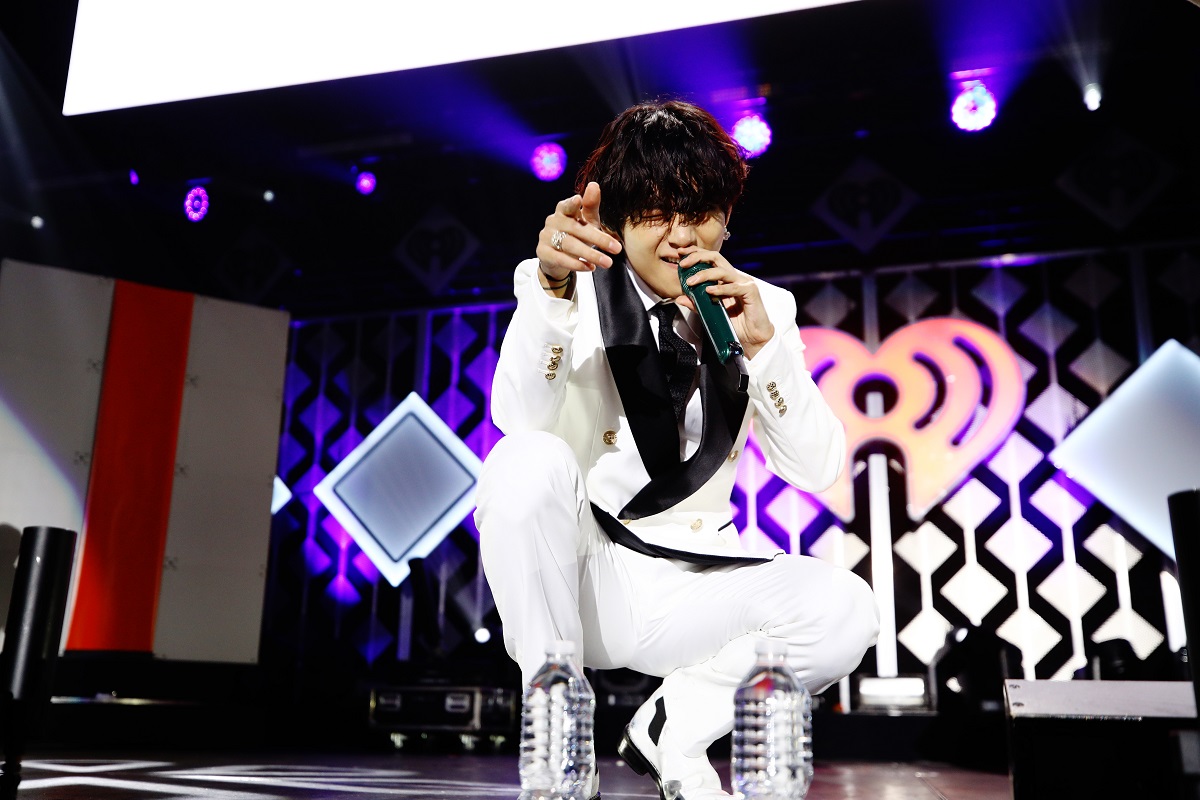 V de la BTS îmbrăcat într-un costum alb cu revere negre în timp ce cântă la microfon și gesticulează